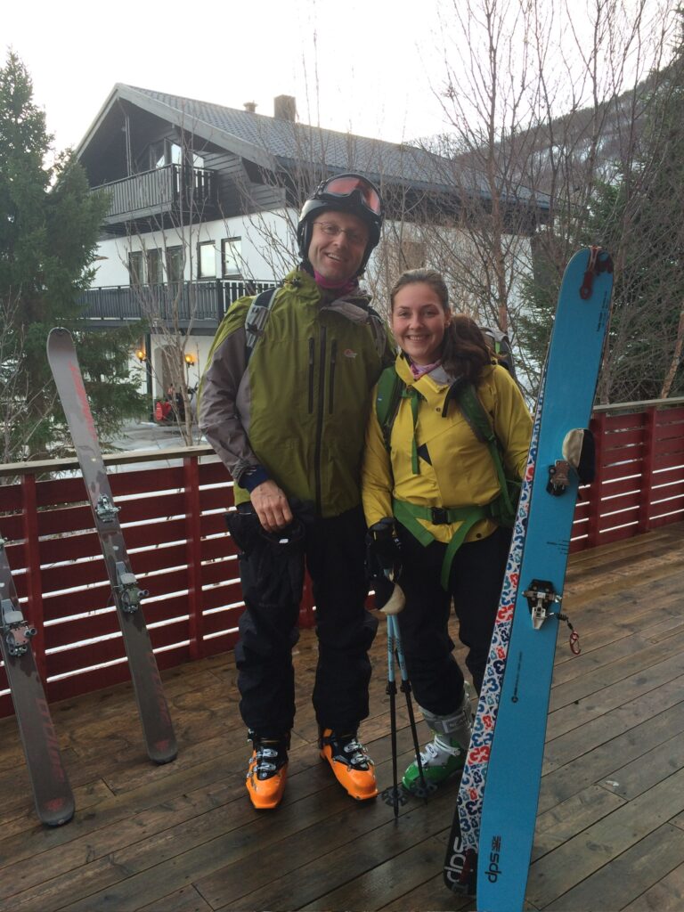 To personer i skiutstyr står på et tredekke med ski og staver og smiler til kameraet. En bygning og trær er synlige i bakgrunnen.