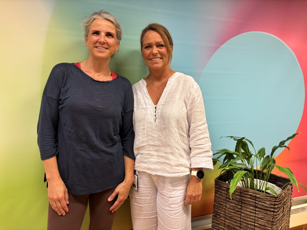 To kvinner står side om side og smiler til kameraet foran en fargerik vegg med en potteplante i nærheten.
