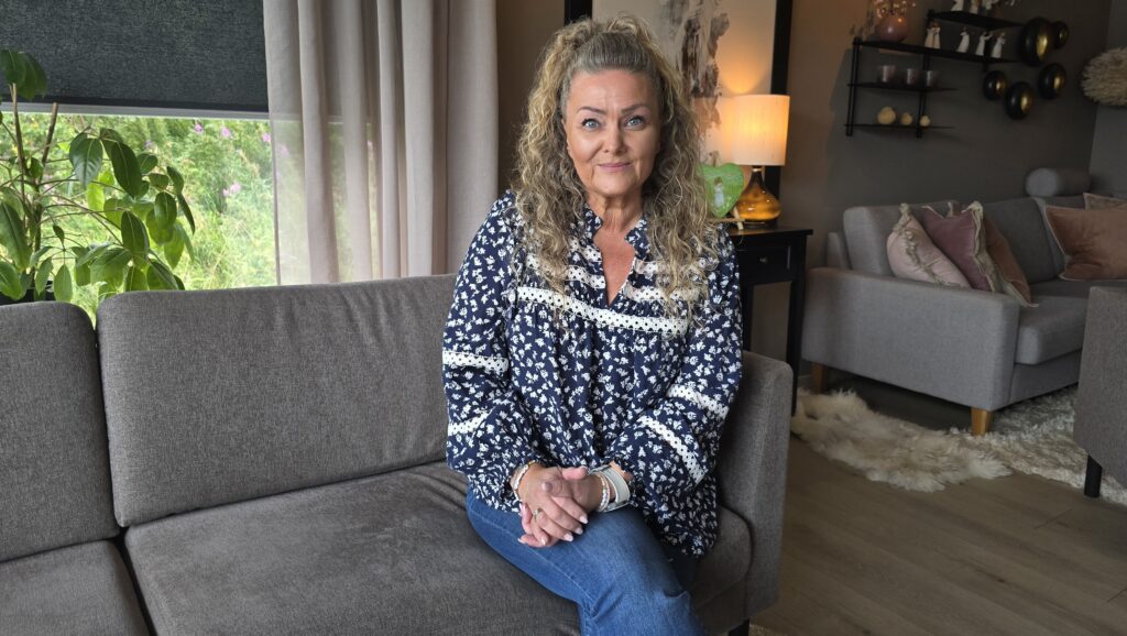 En kvinne med krøllete hår sitter i en grå sofa i en godt opplyst stue med planter og moderne innredning.