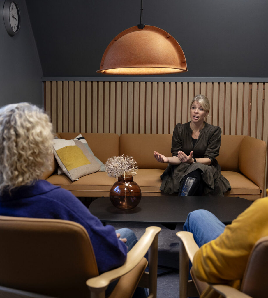 Tre personer sitter i et moderne, svakt opplyst rom med brune møbler; en kvinne snakker, mens to andre lytter overfor henne.