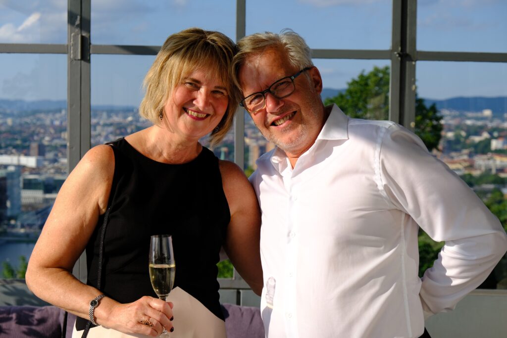 En kvinne med et glass champagne i hånden står ved siden av en mann; begge smiler, og bak dem kan man se et bylandskap gjennom store vinduer.