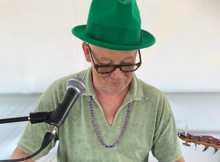 Bak en mikrofon sitter en person i grønn hatt og skjorte, iført briller og et lilla perlekjede, med en gitar delvis synlig ved siden av seg.