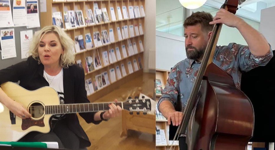 En kvinne spiller akustisk gitar og synger, mens en mann spiller kontrabass; de opptrer innendørs i nærheten av hyller fylt med brosjyrer eller bøker.