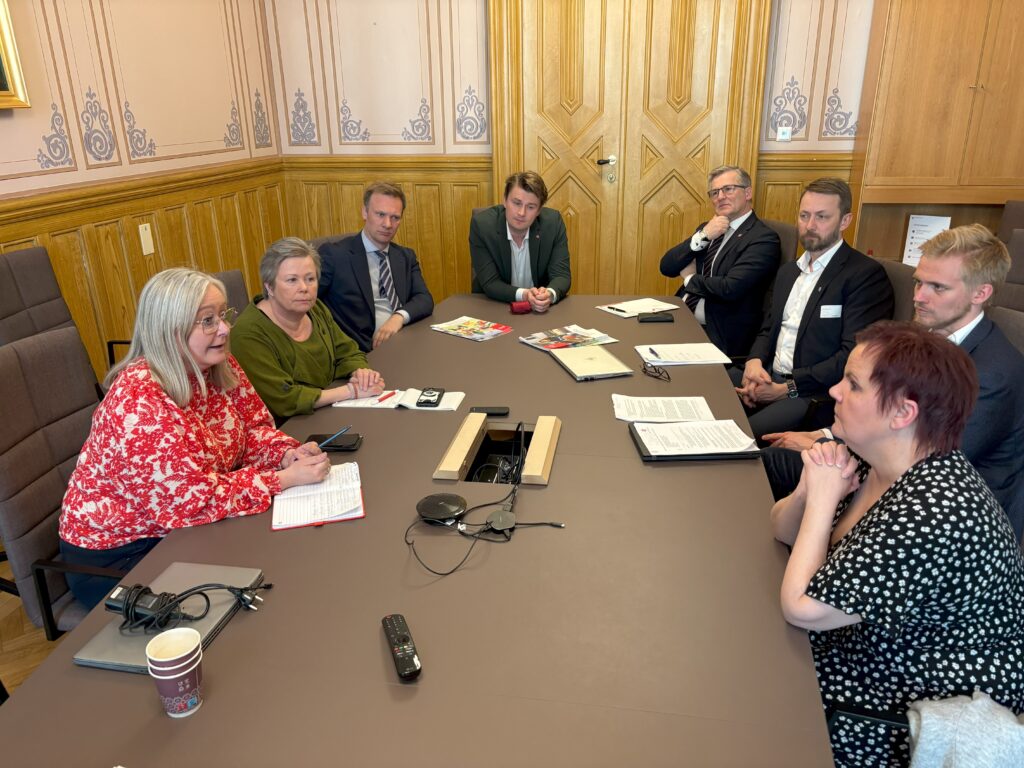 Åtte personer sitter rundt et konferansebord i et rom med trepanel og er i gang med et møte med dokumenter, notatbøker og elektronisk utstyr på bordet.