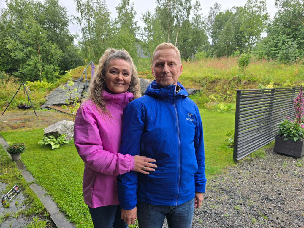 To voksne står utendørs i en hage en overskyet dag, iført regnjakker; kvinnen er i rosa og mannen er i blått. I bakgrunnen er det grønne planter og en huske.