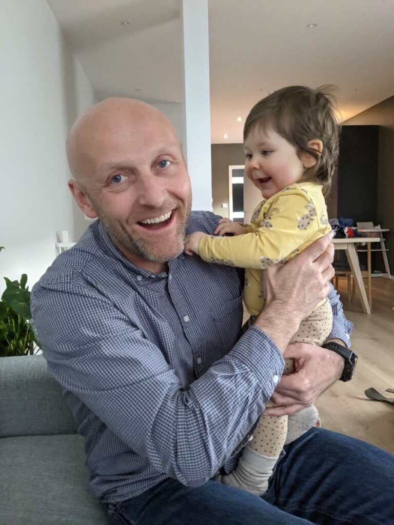 En mann sitter i en sofa og smiler, mens han holder rundt et lite barn som også smiler. De befinner seg innendørs i en moderne stue.