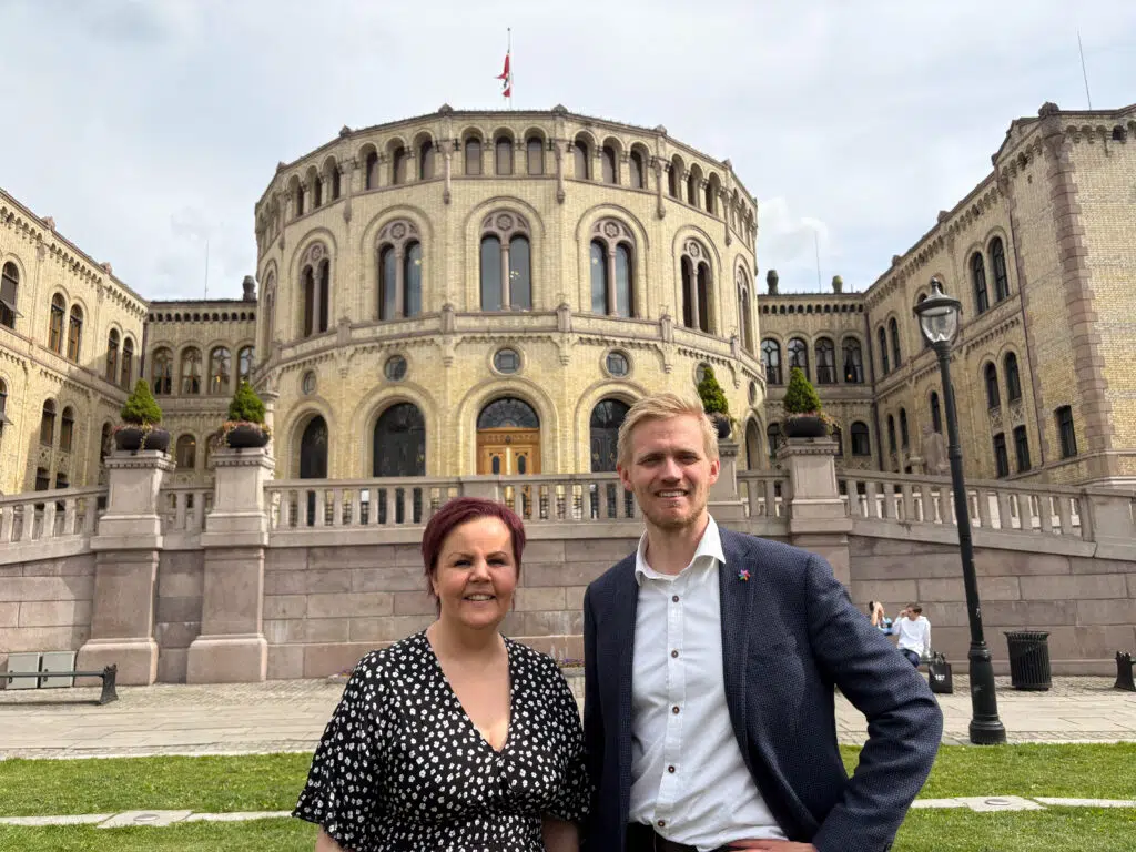 To personer står og smiler foran Stortinget på en overskyet dag.