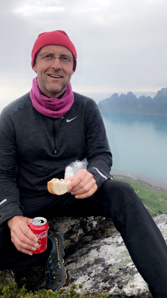 En mann i friluftsklær sitter på en stein med en sandwich og en boks Coca-Cola i hånden, med en innsjø og fjell i bakgrunnen.