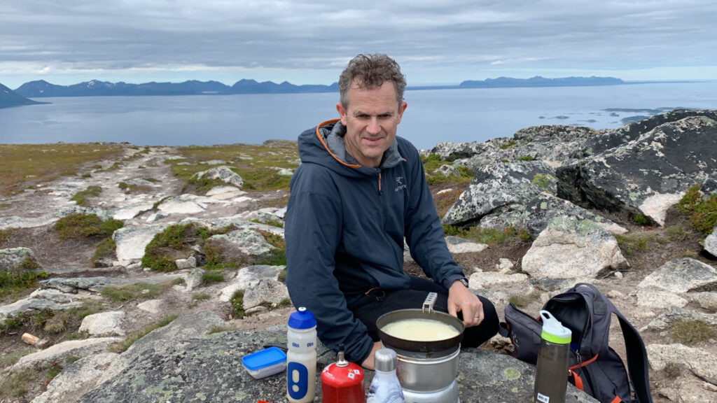 En mann sitter i steinete terreng med campingutstyr og matlagingsutstyr og tilbereder mat utendørs i nærheten av et vann under en overskyet himmel.