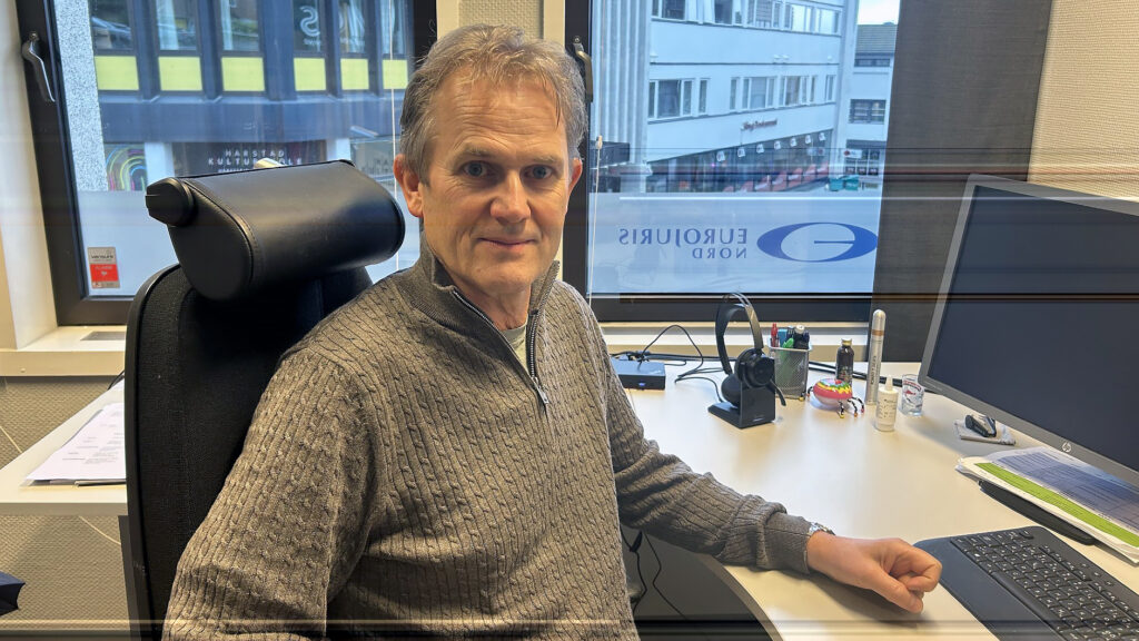 En mann sitter ved et kontorpult med datamaskin, papirer og kontorrekvisita, med vinduer som viser bybygninger i bakgrunnen.