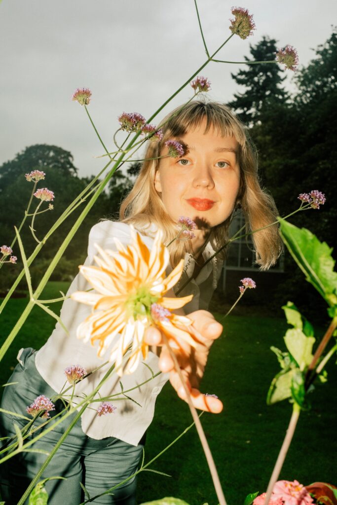 En person med blondt hår og hvit skjorte strekker seg mot en stor gul blomst i en hage, med grønne planter og lilla blomster i bakgrunnen.