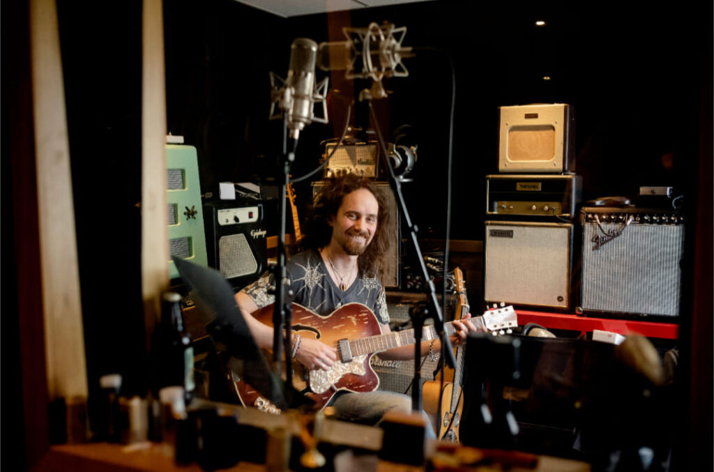En mann sitter i et innspillingsstudio og spiller på en elektrisk gitar, omgitt av forsterkere, mikrofoner og lydutstyr.