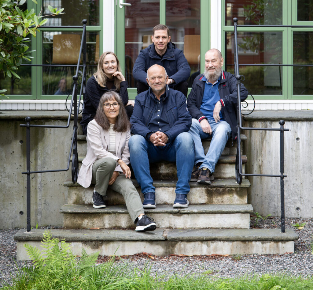 Fem voksne sitter og står på en utendørs trapp foran en moderne bygning med store, grønne vinduer og poserer for et gruppebilde.