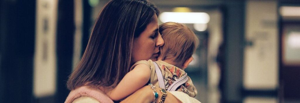 Kvinne holder og trøster et lite barn i en gang, begge vendt bort fra kameraet.