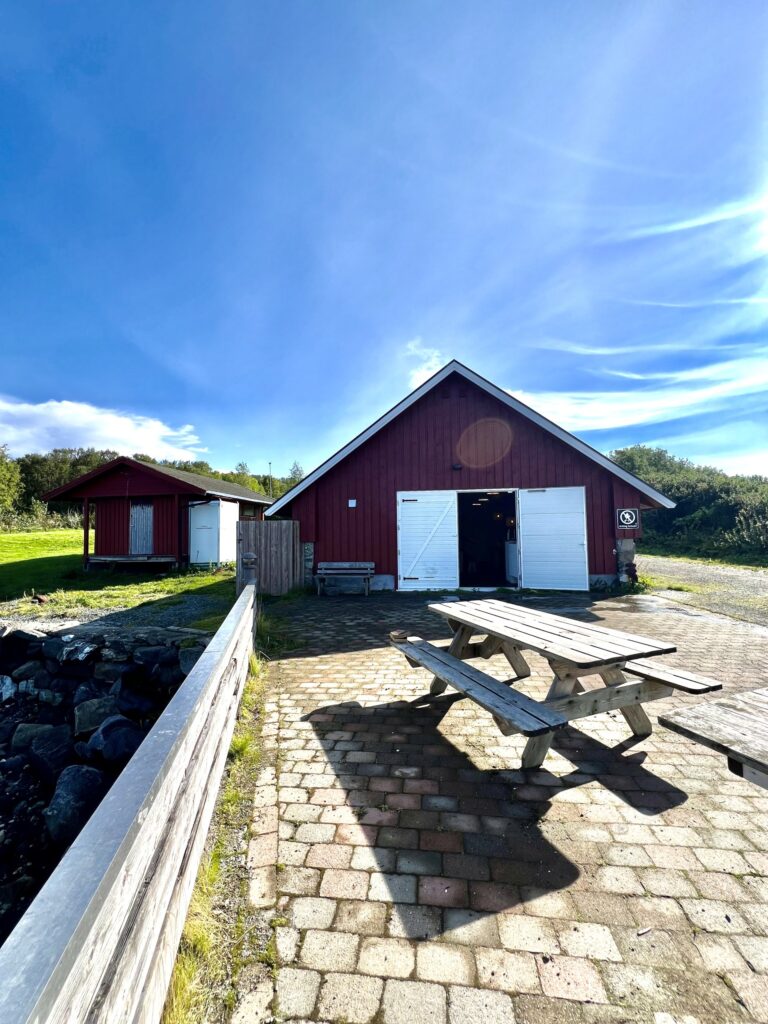 En rød trebygning med hvite dører står under en blå himmel. I forgrunnen står et piknikbord på et steinbelagt område, med gress og en annen liten bygning i nærheten.