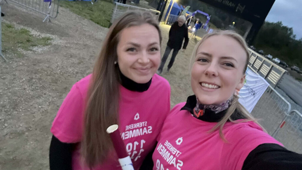 To kvinner i matchende lyserosa T-skjorter smiler når de tar en selfie under et utendørsarrangement med avsperringer og andre deltakere i bakgrunnen.