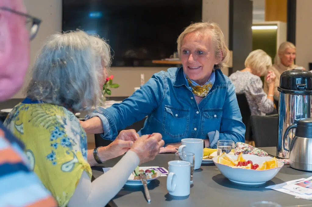 En gruppe eldre voksne sitter rundt et bord med mat og drikke og snakker sammen i lyse omgivelser innendørs.