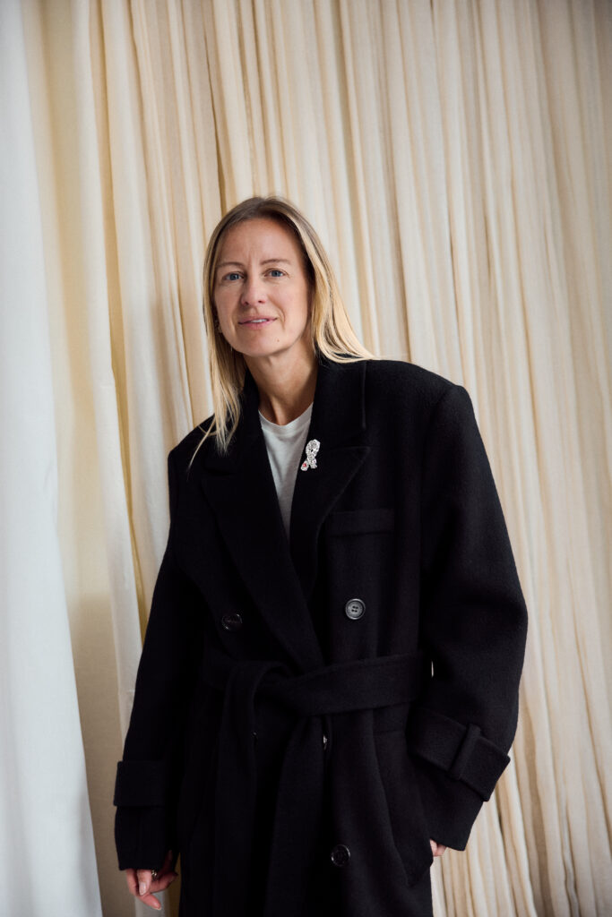 Celine Aagaard med rett blondt hår og en lang svart frakk står foran kremfargede gardiner.