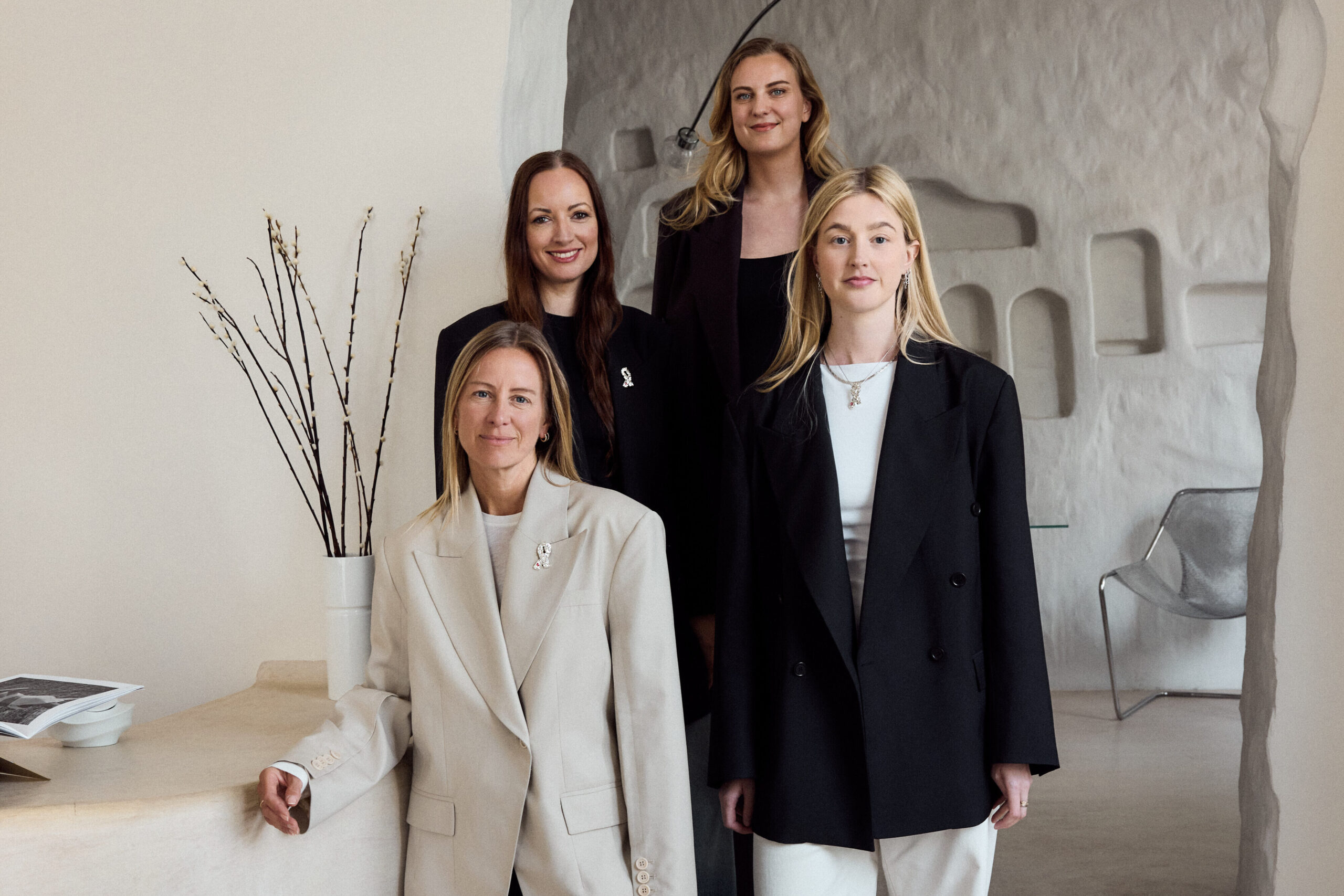 Fire kvinner i blazer og bukse poserer innendørs ved en strukturert buegang og en vase med grener, i et moderne, nøytralt rom.
