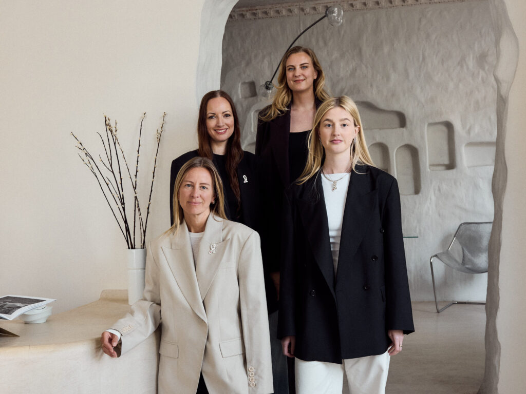 Designteamet i ENVELOPE1976 i blazer og bukse poserer innendørs ved en strukturert buegang og en vase med grener, i et moderne, nøytralt rom.