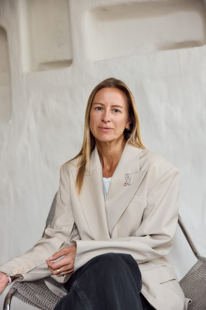 Celine Aagaard med blondt, glatt hår sitter på en stol, iført en beige blazer over en hvit topp og svarte bukser, med en nøytral bakgrunn.