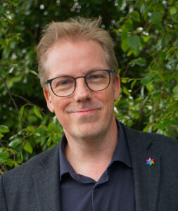 En mann med briller og lyst hår, iført mørk jakke og skjorte, står foran grønne blader og smiler til kameraet.