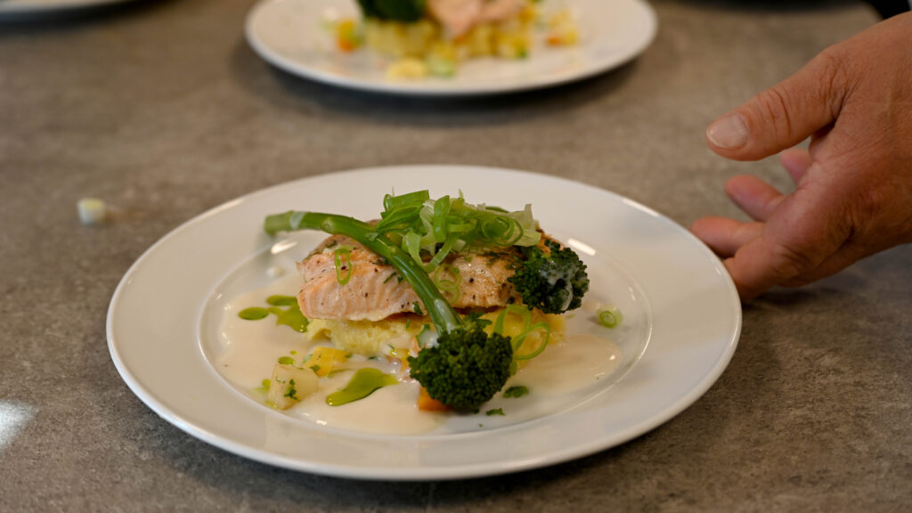 En tallerkenrett med kokt laks på en seng av grønnsaker med brokkoli, overhælt med hvit saus, står på en hvit tallerken på en grå bordplate. En hånd er nær kanten av tallerkenen. Mat, middag, fisk, laks,