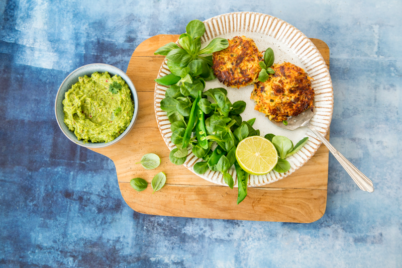 En tallerken med to fritter, friske grønnsaker og en halv lime står på et trebrett ved siden av en bolle med guacamole på blå bakgrunn.