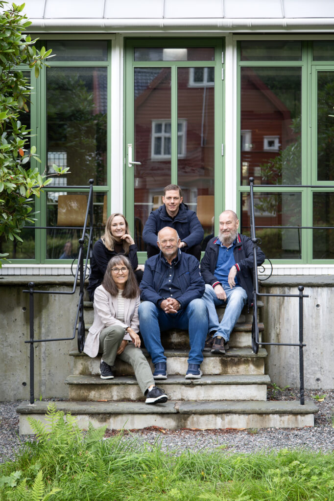 Fem voksne sitter og står på trappetrinn foran en bygning med store, grønne glassdører og vinduer, og poserer for et gruppebilde.