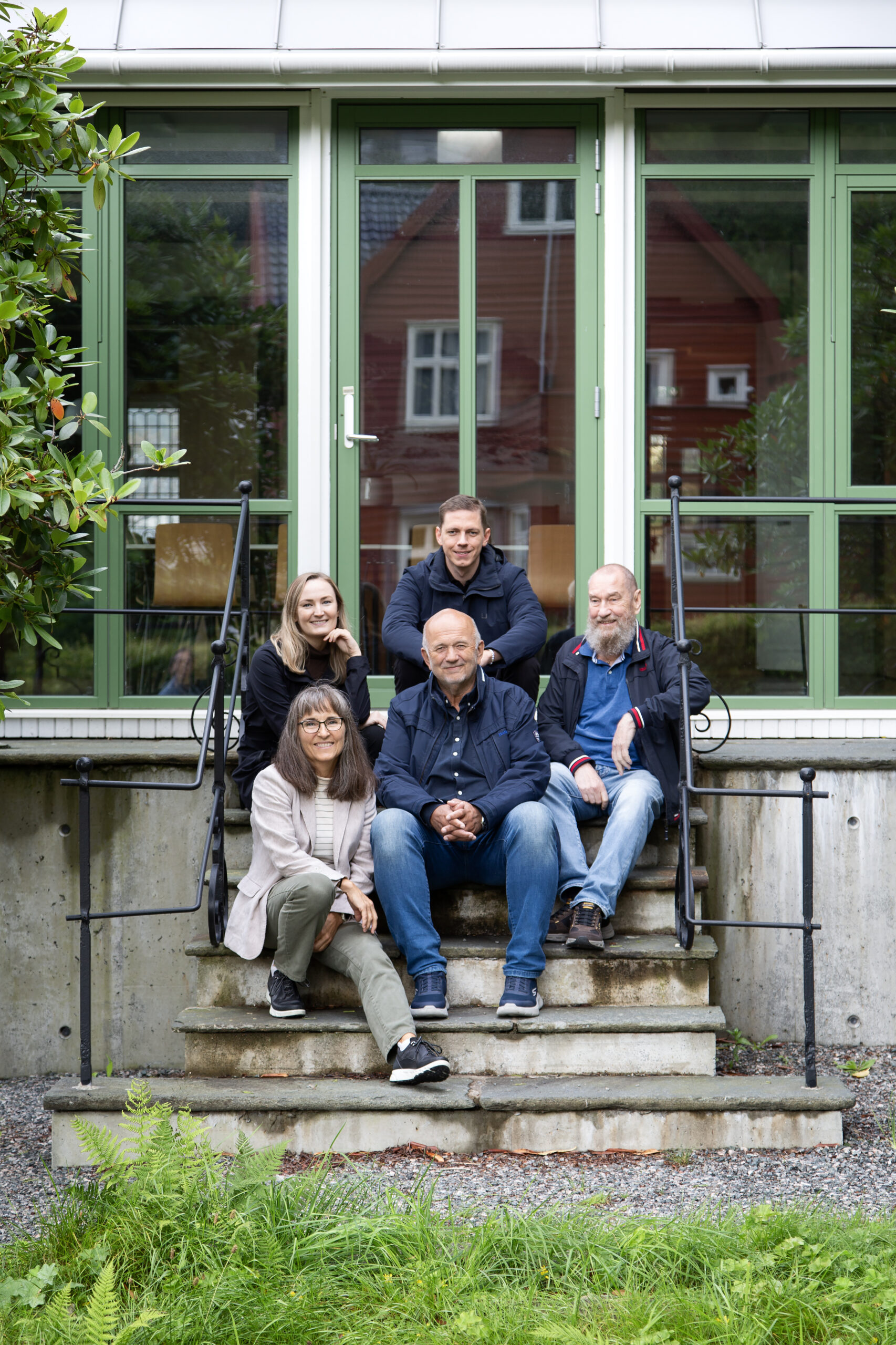 Fem voksne sitter og står på trappetrinn foran en bygning med store, grønne glassdører og vinduer, og poserer for et gruppebilde.