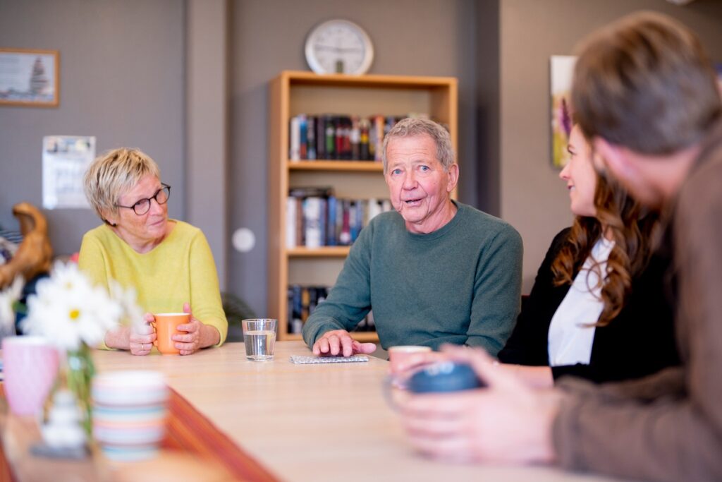 Fire voksne mennesker sitter rundt et bord og fører en samtale i et rom med hyller, bøker og en klokke på veggen. En mann snakker mens de andre lytter.