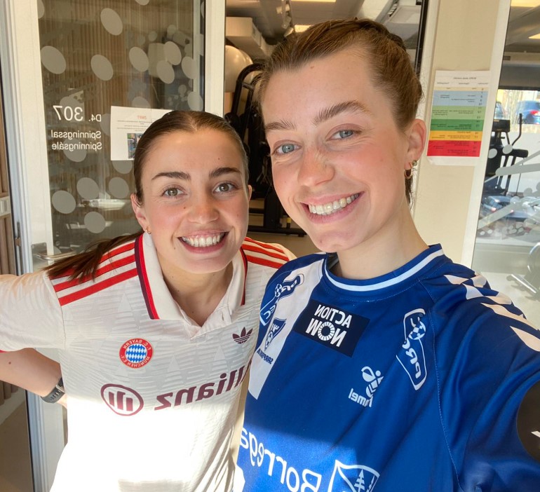 To kvinner i sportstrøyer smiler når de tar en selfie innendørs, side om side i et godt opplyst rom med treningsutstyr i bakgrunnen.