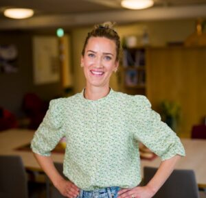 En kvinne med bakoverbøyd hår og en lysegrønn, mønstret bluse står smilende med hendene på hoftene i et innendørs kontormiljø.