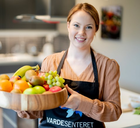 En kvinne på et kjøkken holder en trebolle fylt med diverse frisk frukt mens hun har på seg et svart forkle med påskriften "VARDESENTERET".