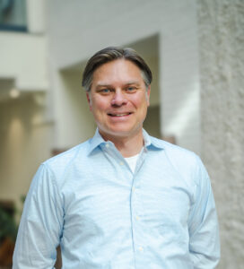 En mann i lyseblå skjorte står innendørs og smiler til kameraet med hendene bak ryggen.