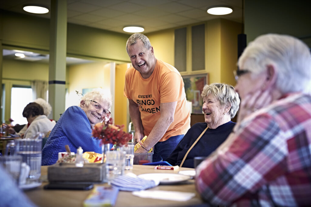 En middelaldrende mann ler sammen med tre eldre damer. Hver fredag spiller de bingo sammen.