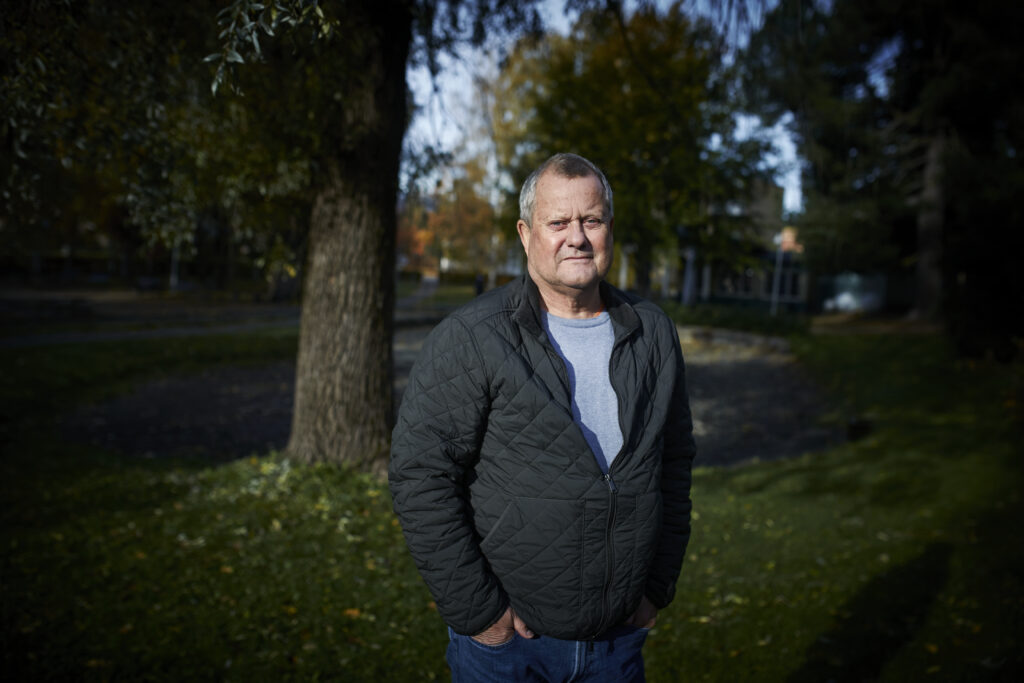 En middelaldrende mann iført mørk vattert jakke står utendørs i en park med trær og gress i bakgrunnen.