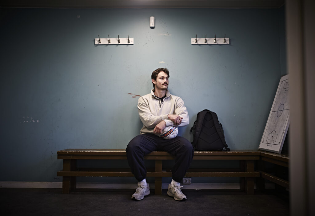 En mann i sportsklær sitter alene på en benk i et garderoberom med en ball i hånden, med en ryggsekk og et strategibrett i nærheten.