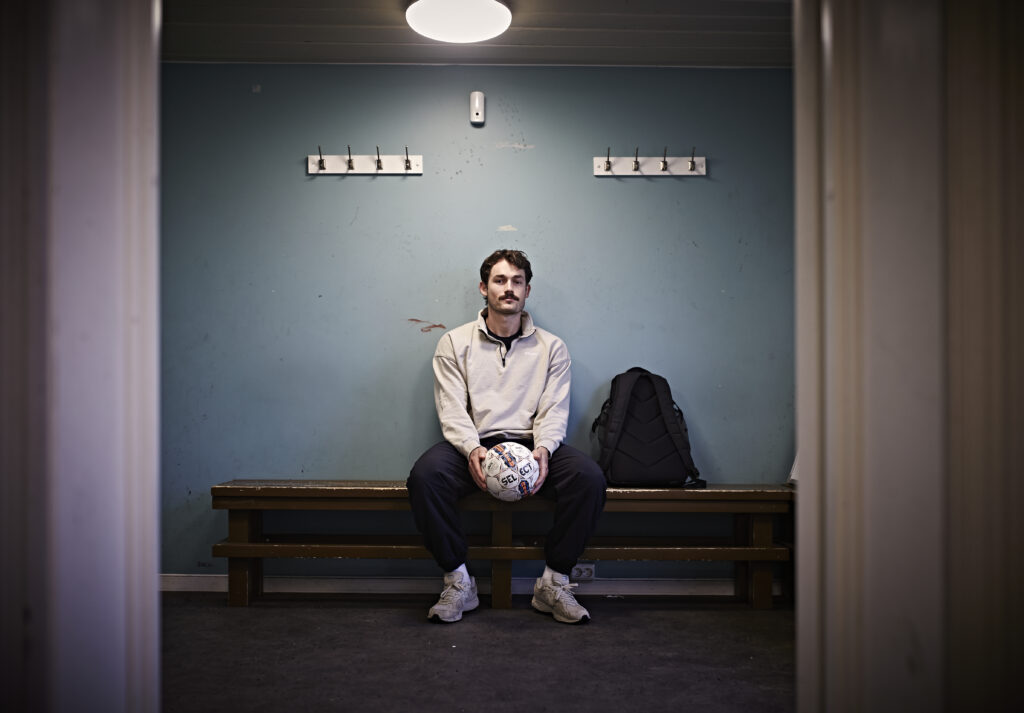 En mann sitter alene på en benk i et garderoberom med en fotball i hånden, med en ryggsekk ved siden av seg og knagger på veggen bak.