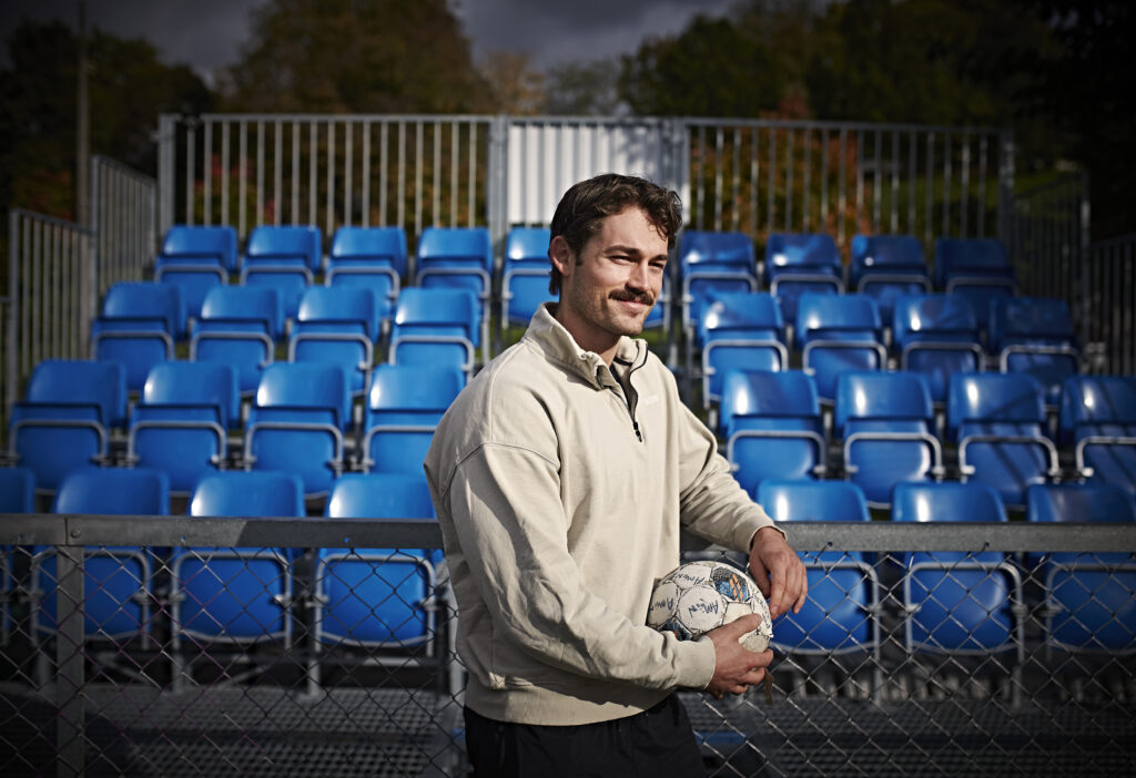 En mann står foran tomme, blå stadionstoler, holder en fotball og hviler armen på et nettinggjerde.