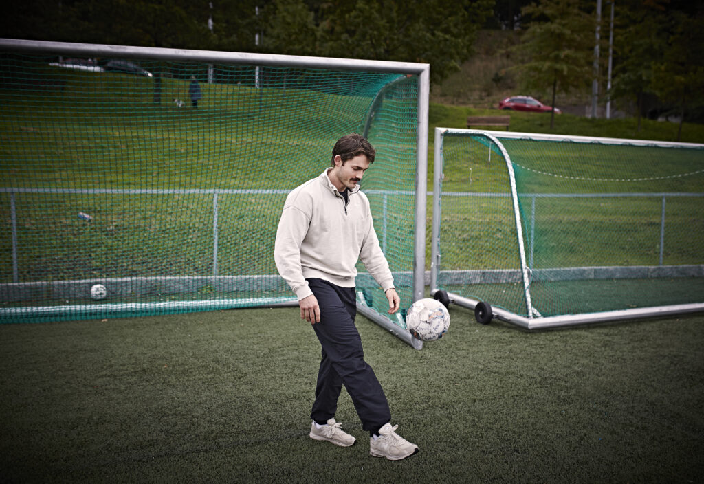 En mann i fritidsklær sparker en fotball mens han står på en utendørs kunstgressbane i nærheten av en målstolpe.