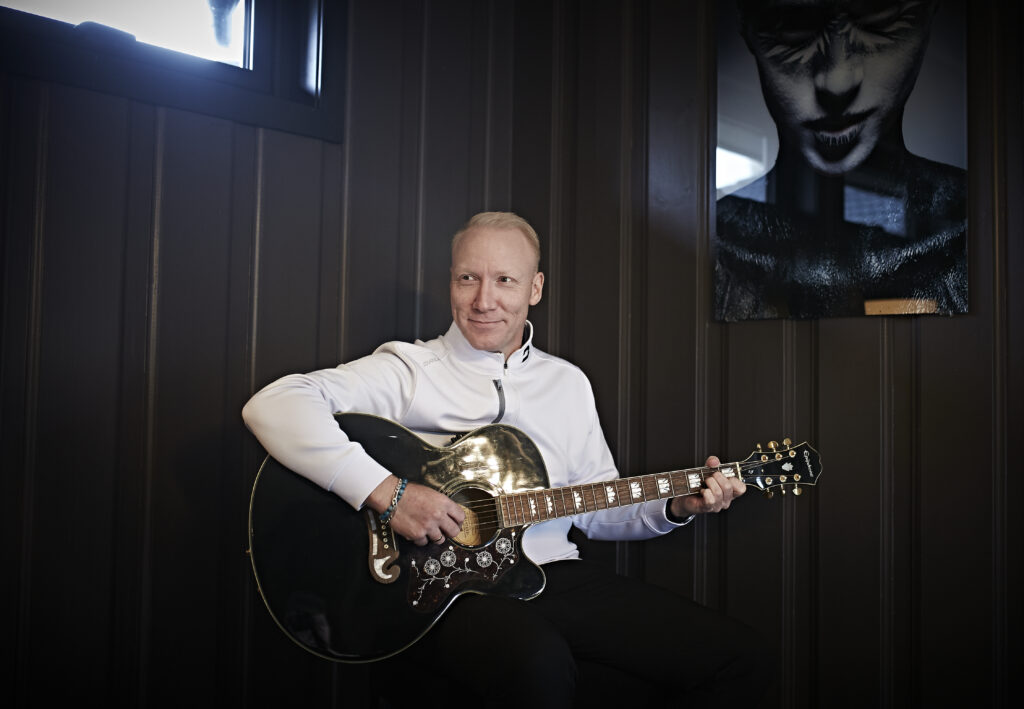 En mann i hvit jakke sitter innendørs og spiller på en svart akustisk gitar. Det er et vindu over ham og et portrettfotografi på veggen ved siden av ham.