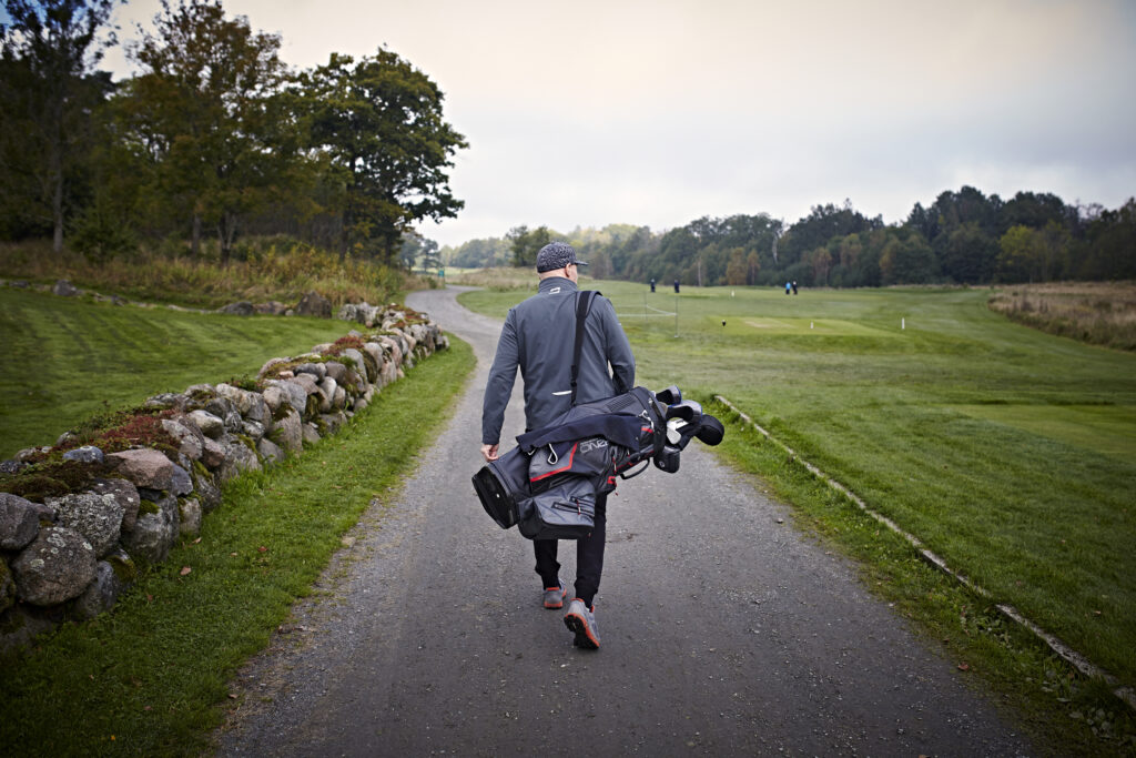 En person med en golfbag går alene på en asfaltert sti mellom gressplener og steinmurer på en golfbane.