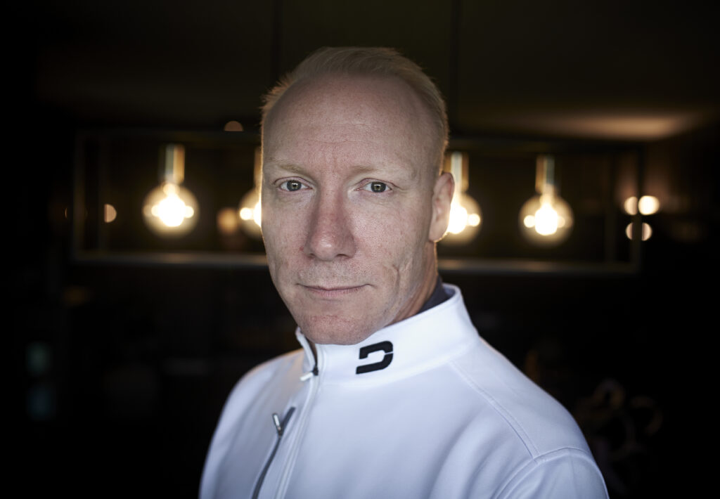 En mann i hvit jakke med glidelås står innendørs med tre hengende lyspærer i bakgrunnen.