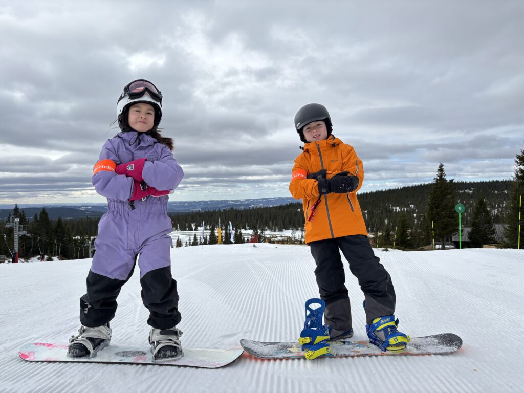 To barn i vinterklær står på snowboard i en preparert skiløype, med trær og en overskyet himmel i bakgrunnen.