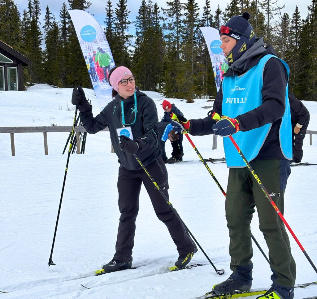 To personer i vintersportsklær står på snøen med langrennsski og staver og snakker med hverandre. Snødekte trær og eventbannere er synlige i bakgrunnen.