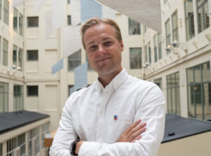 En mann i hvit skjorte står med armene i kors og smiler i et moderne atrium med store vinduer og hvite vegger.