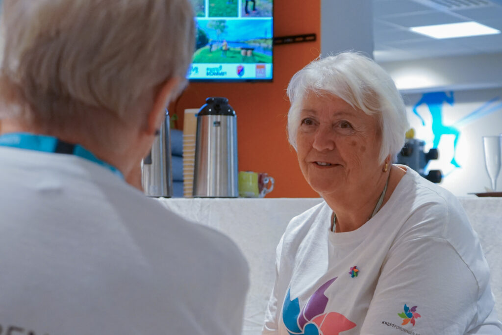 To eldre mennesker i hvite skjorter med fargerike logoer sitter og snakker sammen i et lyst rom med en kaffeautomat og en TV-skjerm i bakgrunnen.