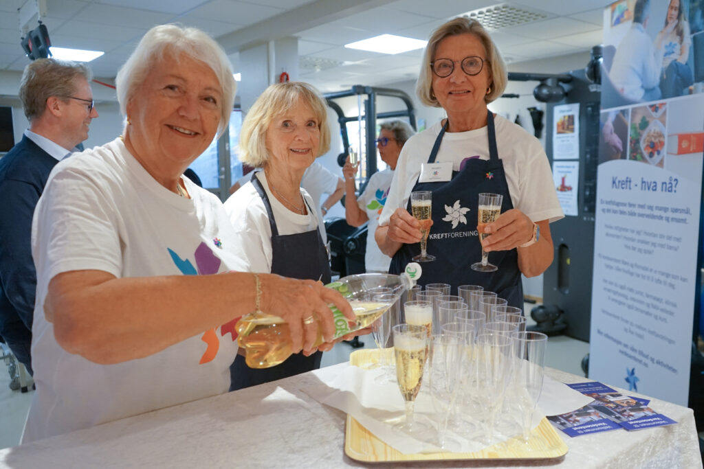 Tre smilende eldre kvinner står bak et bord og skjenker og serverer drinker i plastglass på et innendørsarrangement.