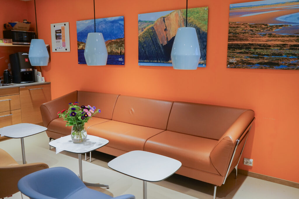 En moderne salong med oransje vegg, brun sofa, små bord, blå pendellamper, veggkunst og en blomstervase på et marmorbord.